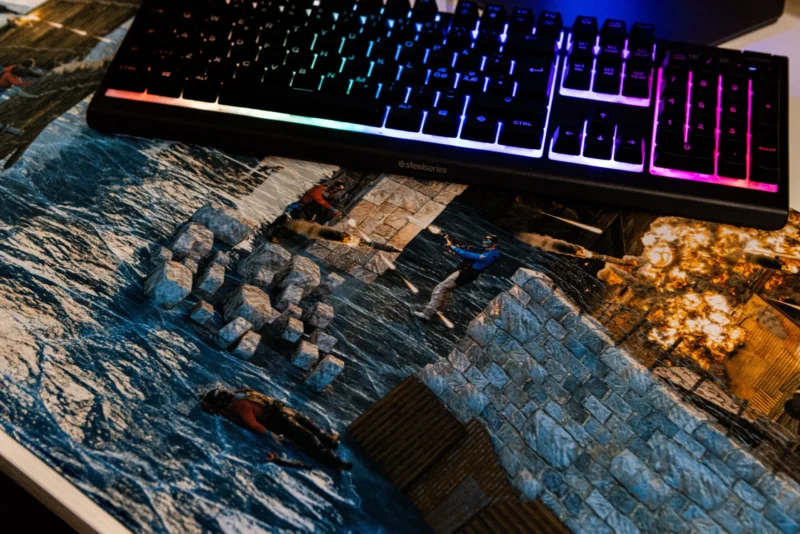 Suur hiirematt lähedalt, millel on kujutatud videomängu Rust lahingut. Hiirematt kaitseb arvutilaua pinda kriimustuste ja kahjustuste eest.